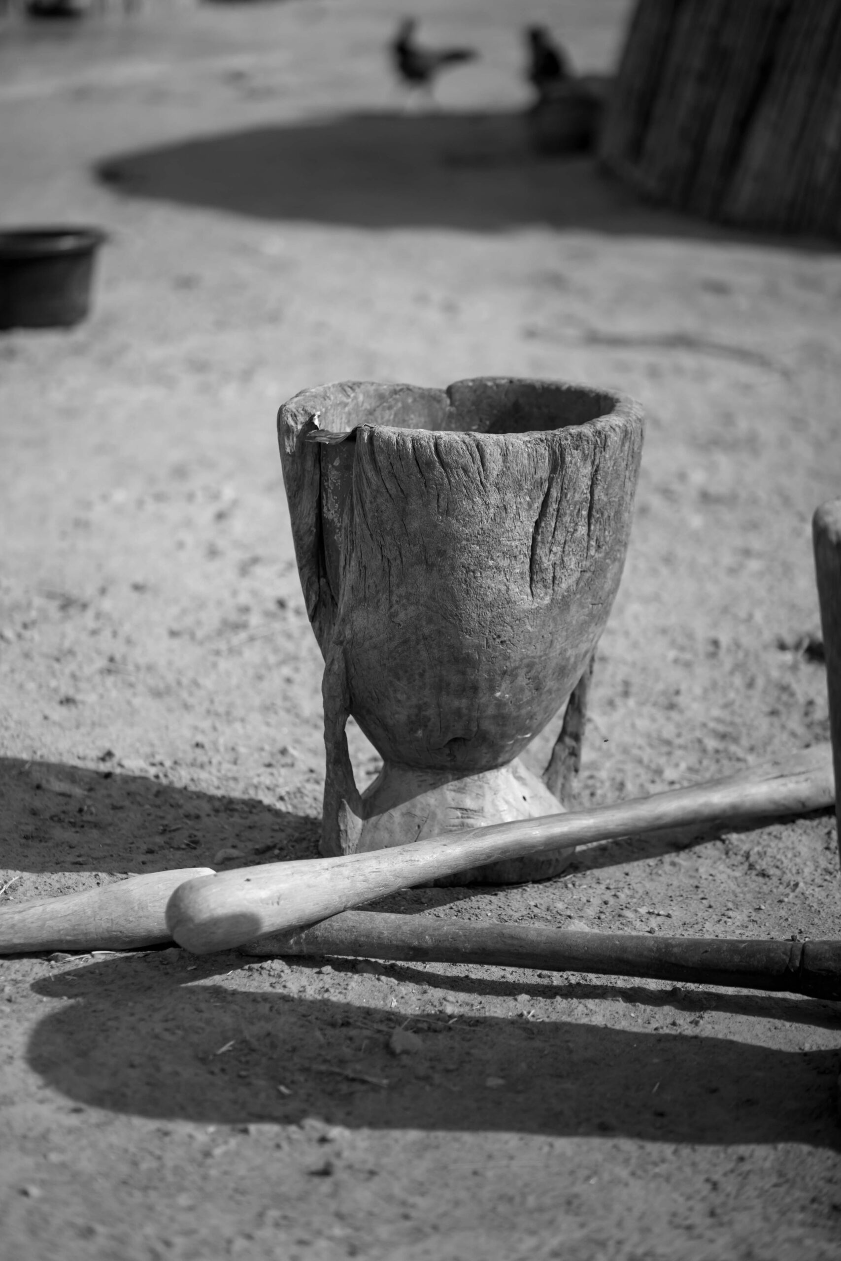 Black and white image of a traditional African wooden mortar and pestle in a rural setting.